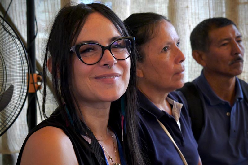 Mujer joven con lentes, sonríe a la cámara. De fondo más participantes de la charla