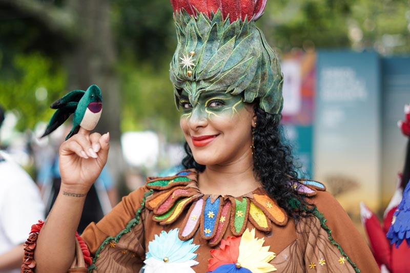 Artista disfrazada con traje verde emulando un árbol, en su mano derecha posa un pájaro de colores