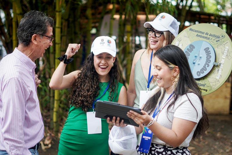 Grupo de tres mujeres compartiendo una sonrisa, con un señor de la tercera edad. Todos celebran mientras interactúan con una tableta, con gorras y distintivos de la COP16 del DNP