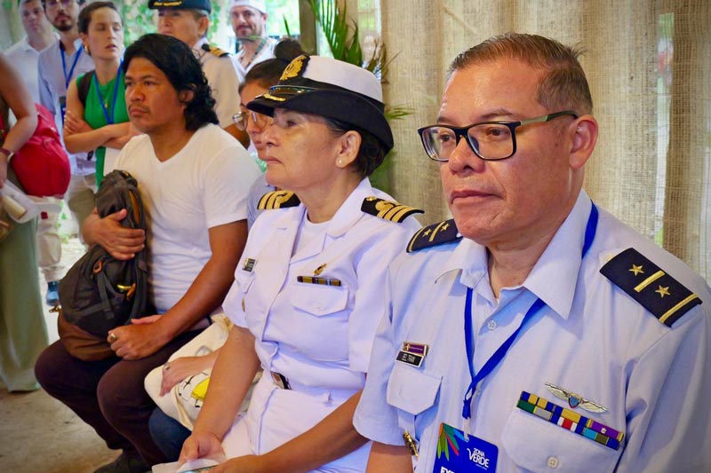 Oficiales navales en uniforme, observando atentamente una presentación en un espacio cerrado. Algunos asistentes al fondo también siguen el evento con interés