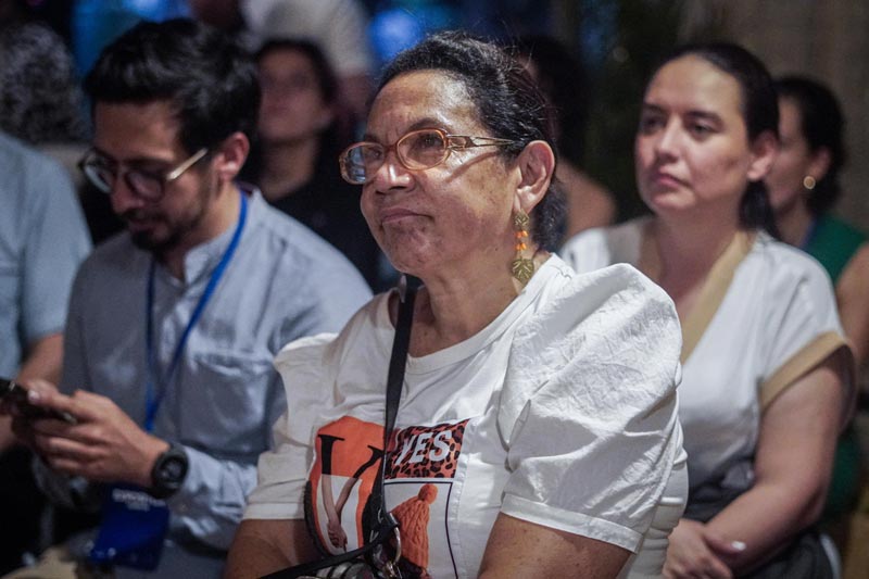 Mujer con gafas y camiseta blanca, junto a otros asistentes, observando atentamente una presentación en un evento. En el fondo, más personas siguen el desarrollo de la actividad