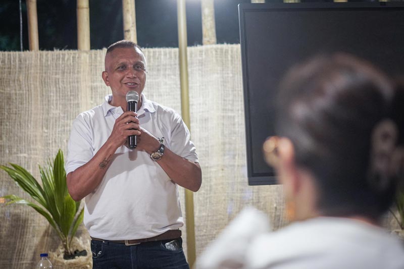 Hombre vestido de camiseta blanca, con micrófono en la mano expone ante la comunidad, temas de biodiversidad.