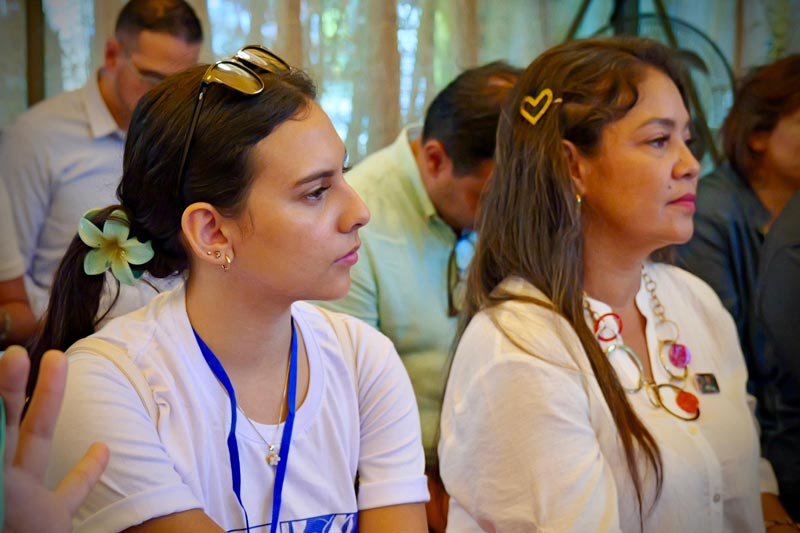 En primer plano, dos mujeres una joven y otra más madura, están atentas a las indicaciones dadas por los conferencistas.