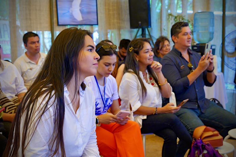 Grupo de personas que escuchan atentamente la conferencia, en primer plano una mujer joven de cabello negro largo.  