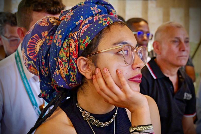 Mujer joven, atenta en la conferencia. Tiene un turbante azul en el cabello y  usa lentes transparentes. 