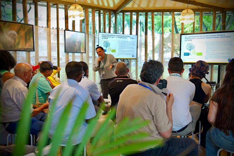 Hombre exponiendo frente a un grupo de personas, en una sala con pantallas y gráficos.  