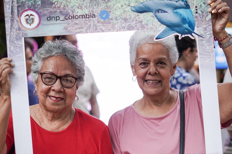 Mujeres adultas mayores, sostiene marco del DNP, decorado con animales marinos.