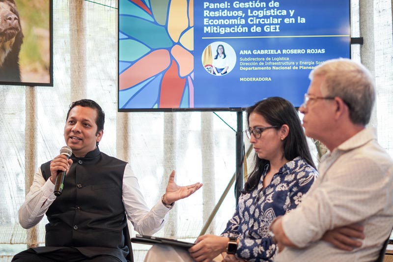 Tres panelistas, dos hombres, una mujer. De fondo una presentación proyectada en una pantalla led.  