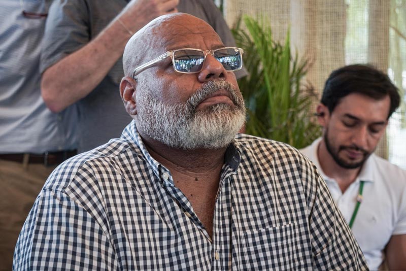 Hombre con camisa de cuadros y gafas transparentes observando atentamente una presentación en un evento, acompañado de otros participantes en el fondo.