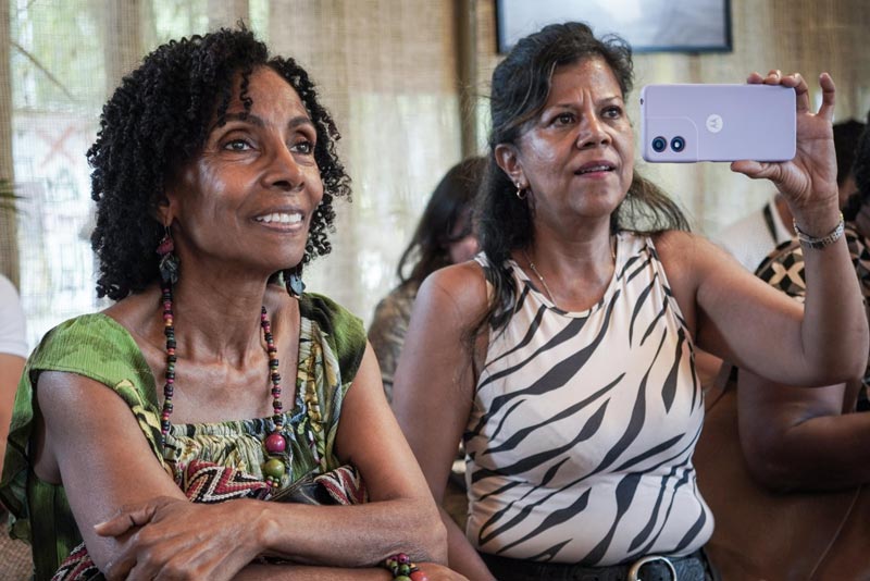 Dos mujeres afro, una de ellas se ve feliz escuchando la charla, mientras la otra está grabando lo que dice el conferencista.  