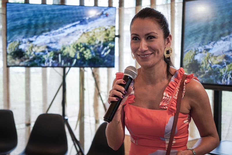 Mujer conferencista, tiene micrófono en mano y se ve feliz. 