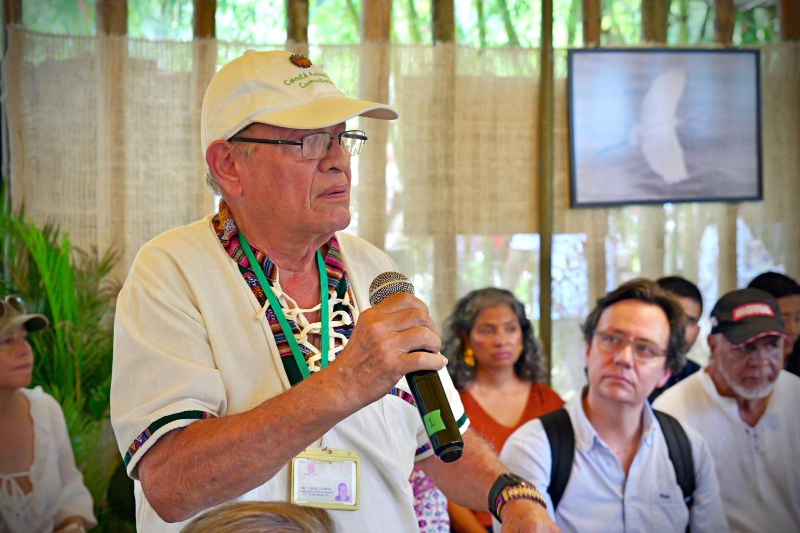 Hombre adulto mayor con gafas y gorra del DNP participa en conversatorio. De fondo más público.