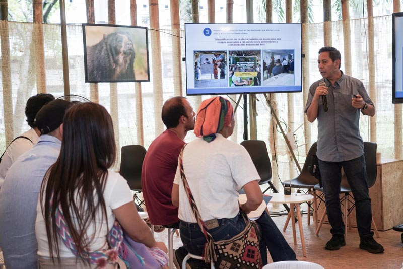 Seis panelistas exponen ante el auditorio lleno, en los laterales hay dos pantallas planas con imágenes ambientales.