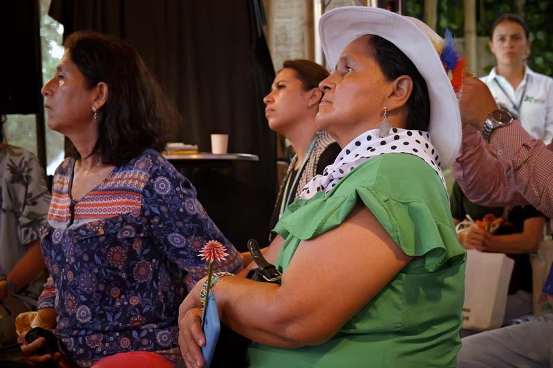 Tres mujeres de edad media, en primer plano con sombrero y blusa verde, concentrada en la charla.