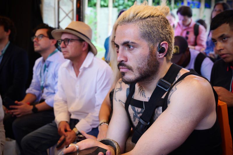 En primer plano, hombre joven de aspecto casual, concentrado viendo la charla. De fondo varios asistentes atentos.  