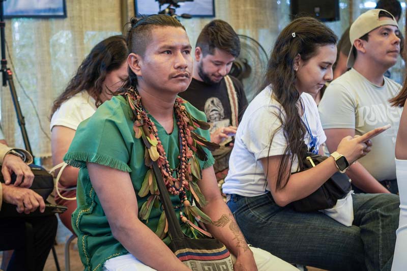 Joven indígena, viste camiseta verde, collar étnico.El junto a varias personas están concentrados en la charla.