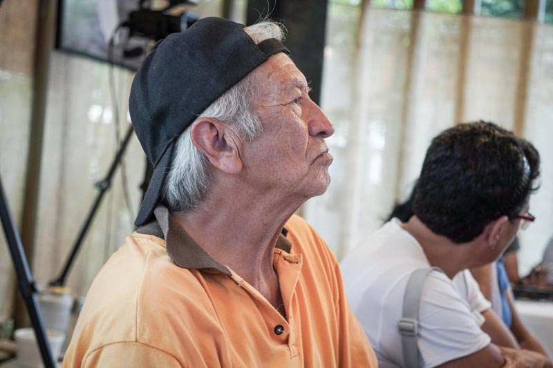 Adulto mayor, con gorra negra hacia atrás y camisa naranja, está concentrado en la charla.  