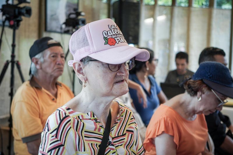 Adulta mayor, viste camisa con rayas de colores, gorra rosada y lentes. Está concentrada en la charla. 