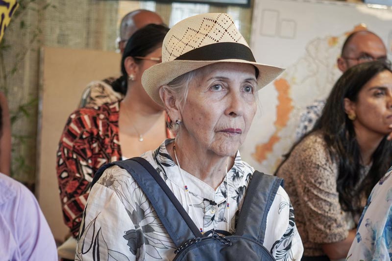 Adulta mayor, con sombrero y morral de frente, está atenta a la charla