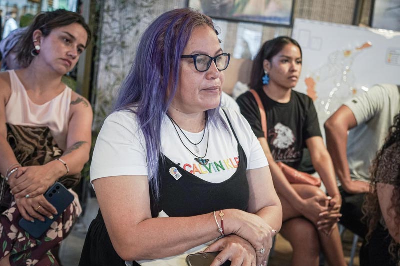 Mujer de mediana edad, cabello morado y lentes, está concentrada en la charla. De fondo publico expectante