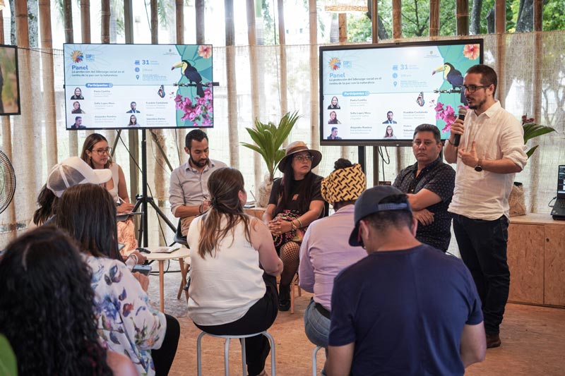 Auditorio lleno, en el panel cinco expertos, presentan imágenes en pantallas laterales. 