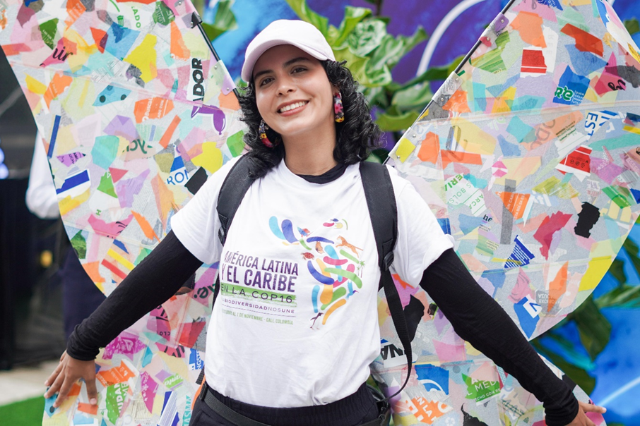 Mujer joven viste camiseta de la COP16. De fondo una mariposa hecha de reciclaje.